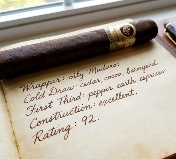 A premium dark Maduro cigar resting foot-out on an open leather notebook of handwritten tasting notes, with a crystal water tumbler and a brass reading lamp on a mahogany side table.