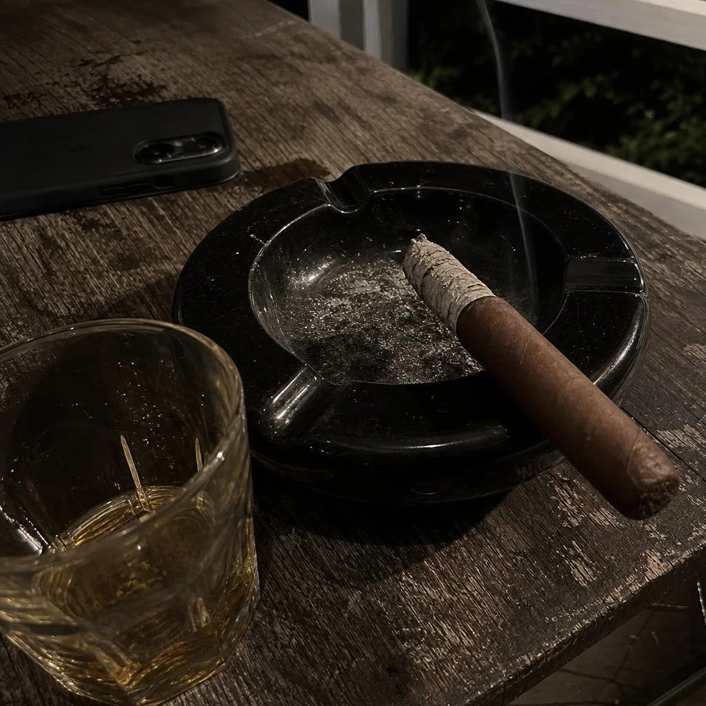 A lit robusto in a black ceramic ashtray with an inch of pale grey ash, seen from above on a rainy porch table
