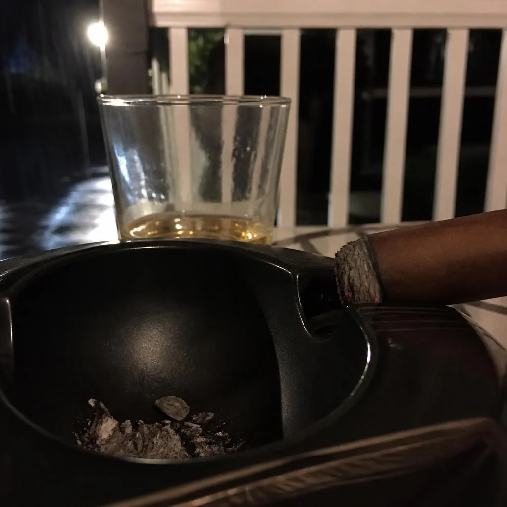 The foot of a lit robusto just after the ash fell, resting in a black ceramic ashtray beside a tumbler on a rainy porch