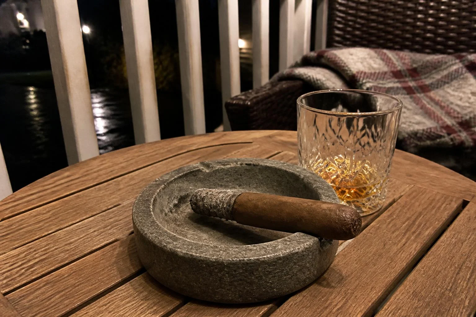 A cigar smoked to its last third resting in a grey stone ashtray beside a crystal tumbler on a wet porch table at night