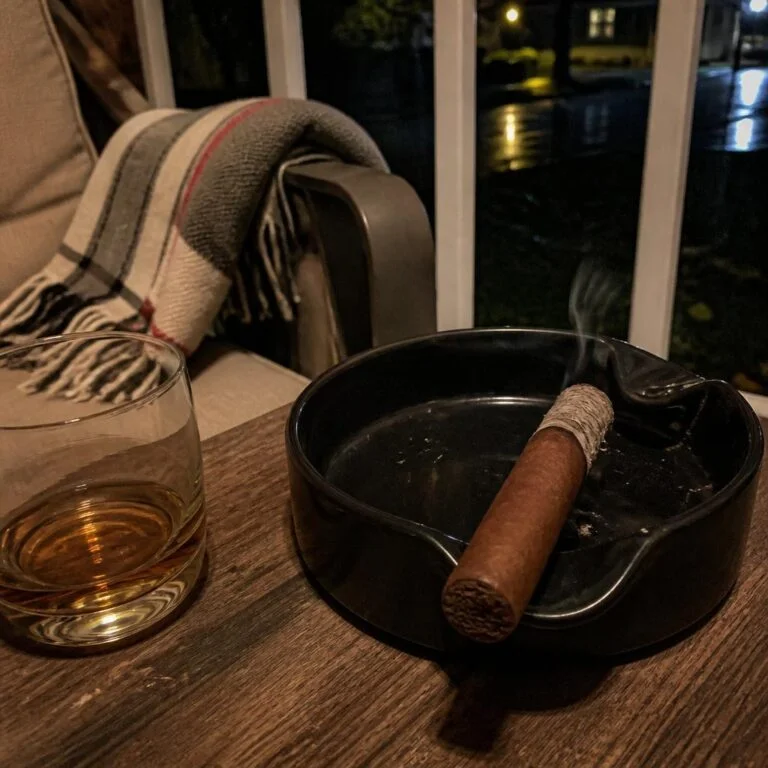 A lit robusto resting in a black ceramic ashtray on a porch side table beside a tumbler of whisky on a wet October night
