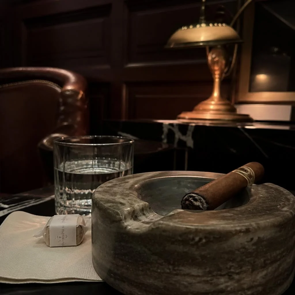 A partly smoked cigar resting in the notch of a heavy stone ashtray beside a glass of water and a wrapped sugar cube on a folded napkin, warm lamplight behind.