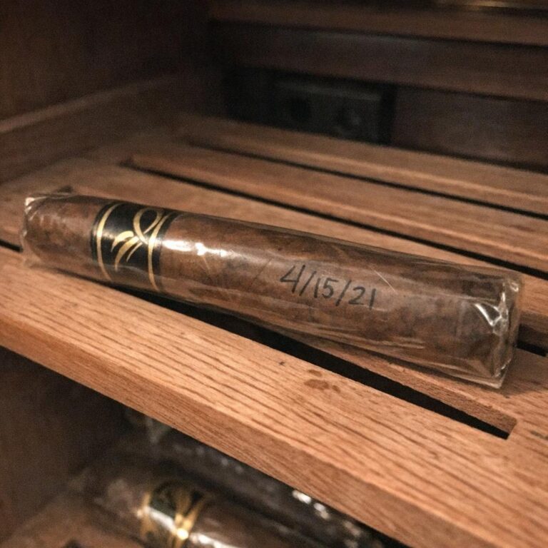 Cellophaned cigar with handwritten date directly on the cellophane sleeve resting on a cedar humidor shelf, brass humidor visible above