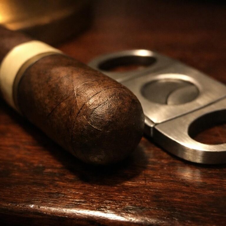 Close-up of an unlit cigar's rounded cap on dark wood beside a steel double-guillotine cutter in warm lamplight.