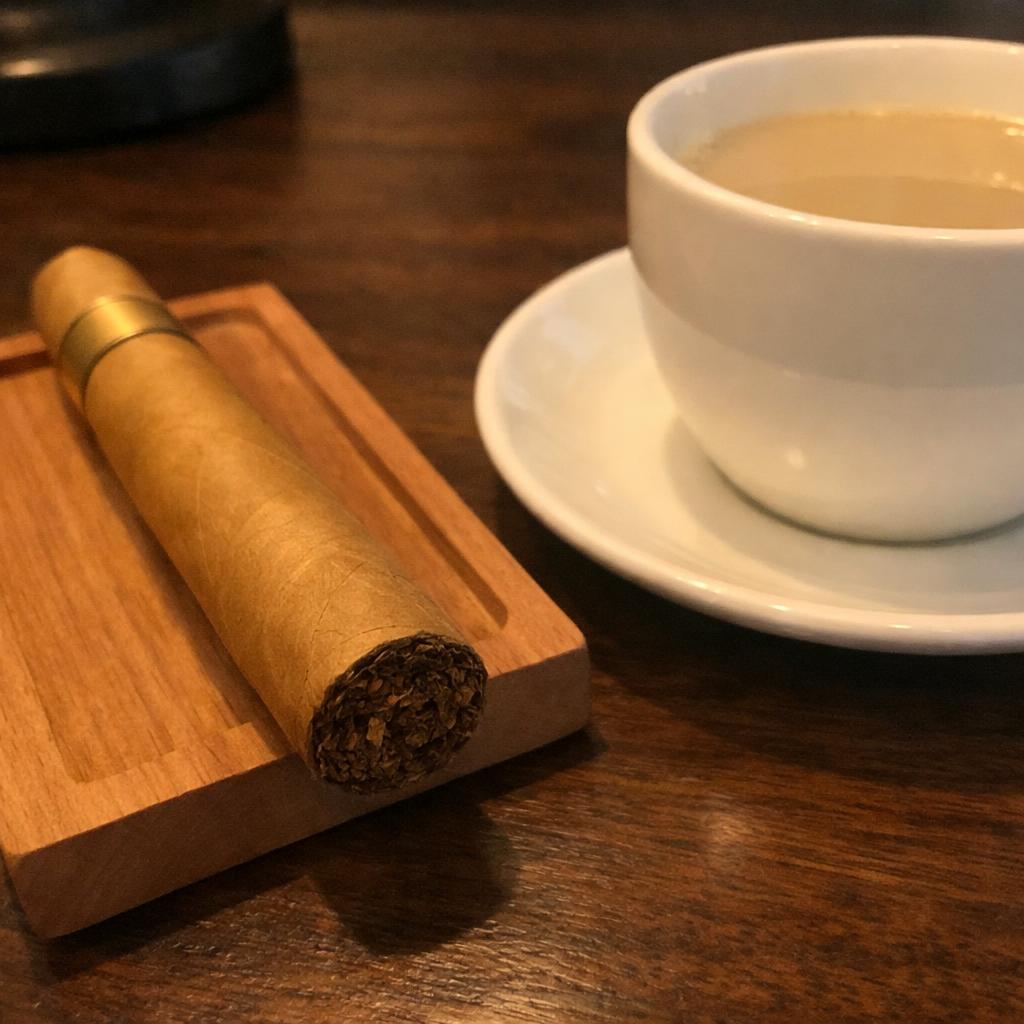 A pale Connecticut-shade cigar rests on a cedar tray beside a cup of light blonde-roast coffee on a dark walnut table.