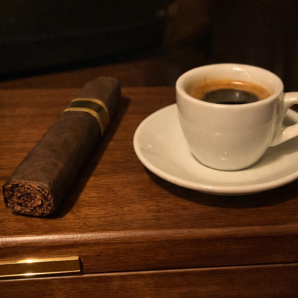 A box-pressed dark maduro cigar rests on a walnut humidor lid beside a small cup of espresso with crema in low light.