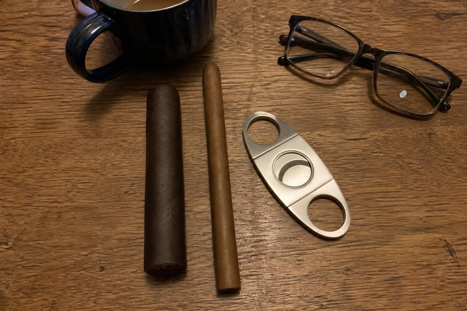 A thick unlit 60 ring gordo and a slim 38 ring lancero lying side by side on a kitchen table beside an open double-blade cutter, a mug of tea and reading glasses