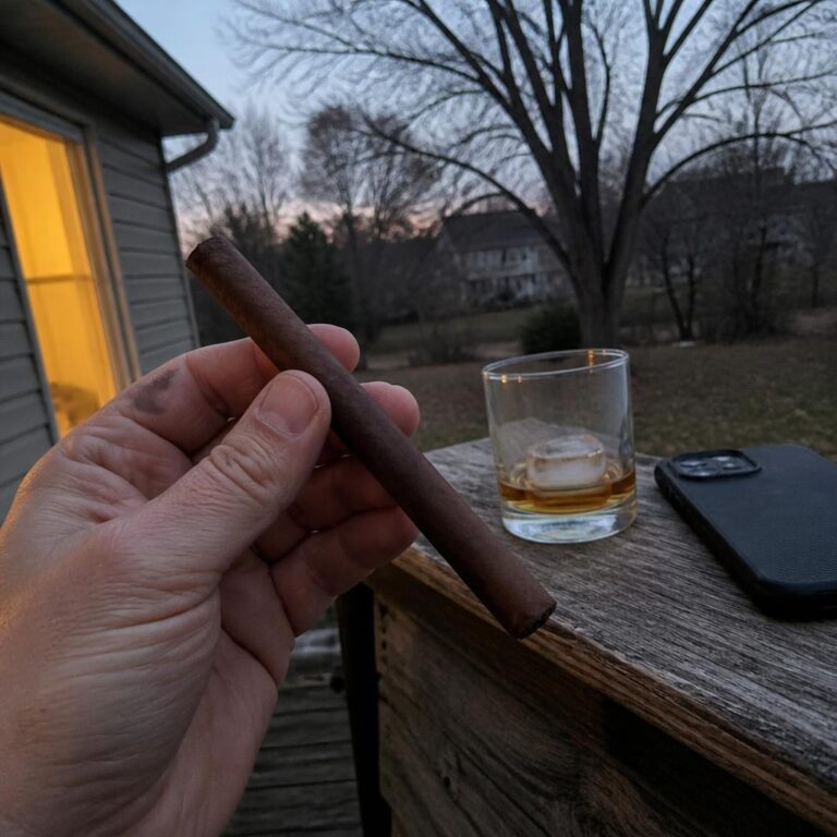 A hand holding a slim unlit corona cigar on a back deck at dusk, beside a half-finished whisky and a phone on the rail