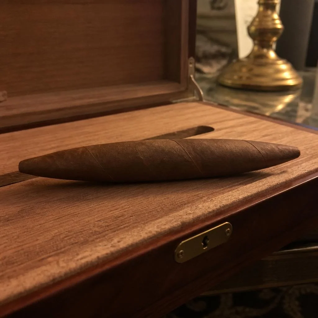 An unlit perfecto cigar with a closed tapered foot resting on the cedar tray of an open humidor beside a brass lamp