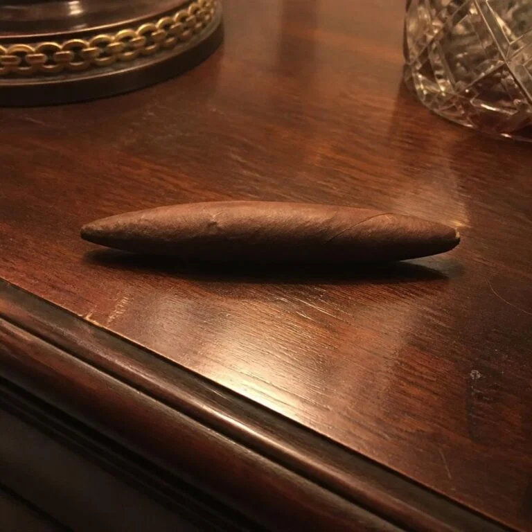 A single unlit perfecto cigar, tapered and closed at both ends, resting on a polished walnut side table in warm lamplight